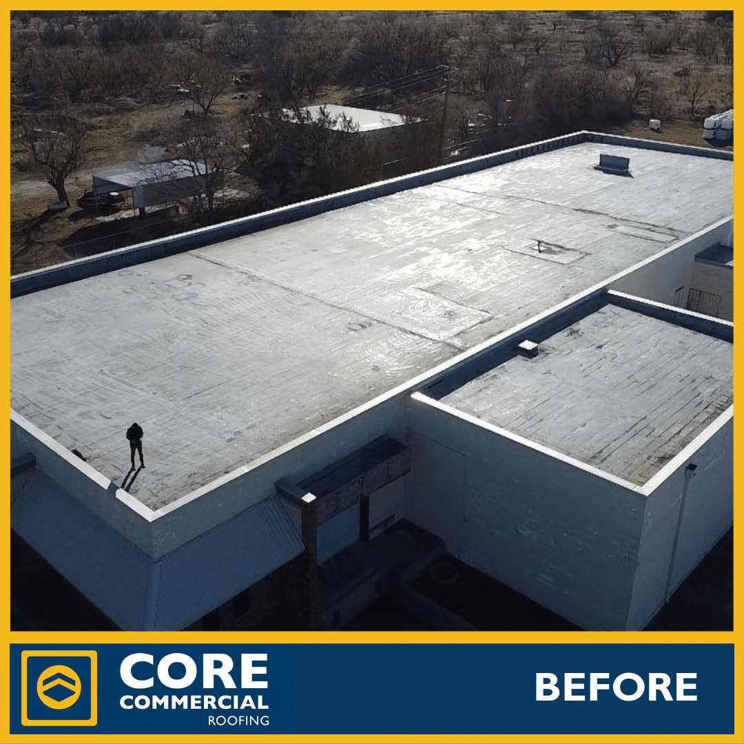 Aerial “before” photo of a large commercial flat roof in poor condition. The roof is covered with an old, heavily stained, and patchy white coating that has failed in large sections, exposing dark substrate. Visible cracks, blisters, open seams, rust stains, and debris. A lone worker stands on the roof for scale. Taken by Core Commercial Roofing in a rural/industrial setting.