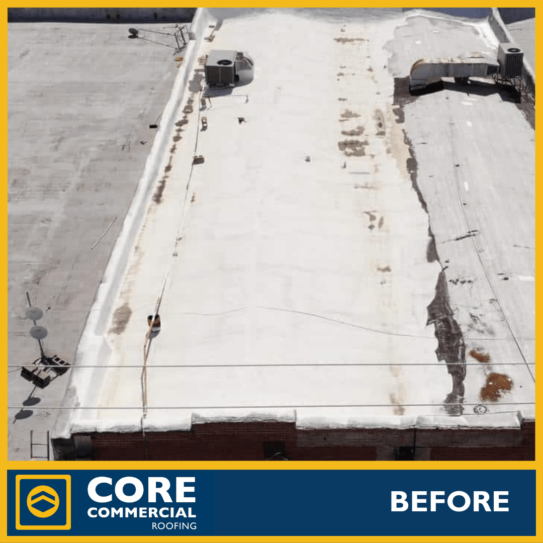 Aerial “before” photo of a large commercial flat roof in poor condition. The roof is coated with an old, failing white reflective coating that has severely stained, discolored, and peeled in large sections, exposing the dark substrate. Heavy rust stains, ponding water marks, open seams, and debris, and multiple roof penetrations are visible. Taken by Core Commercial Roofing.
